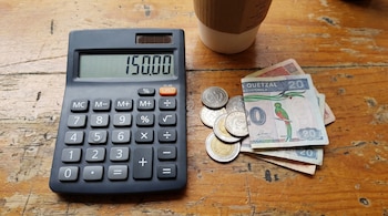Una calculadora negra con el número '150.00' en su pantalla, varias monedas y billetes de Quetzales guatemaltecos y una taza de café sobre una mesa de madera.