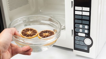 Mano sosteniendo un cuenco de vidrio transparente con agua y dos rodajas de limón secas, introduciéndolo en un horno microondas blanco.