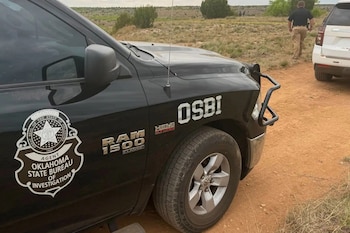 Parte delantera de una camioneta negra de la Oficina de Investigaciones del Estado de Oklahoma (OSBI) con emblema, estacionada en un camino de tierra. Un oficial camina en el fondo