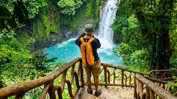 Hombre de espaldas con mochila naranja y sombrero camuflado en sendero de madera. Cascada y río turquesa rodeados de vegetación densa.
