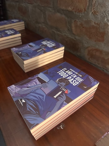Pilas de libros con portada azul y la ilustración de un joven leyendo descansan sobre una mesa de madera junto a una pared de ladrillo