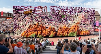 Una carroza decorada con flores moradas y naranjas frente a una multitud de personas sentadas en gradas bajo un cielo despejado