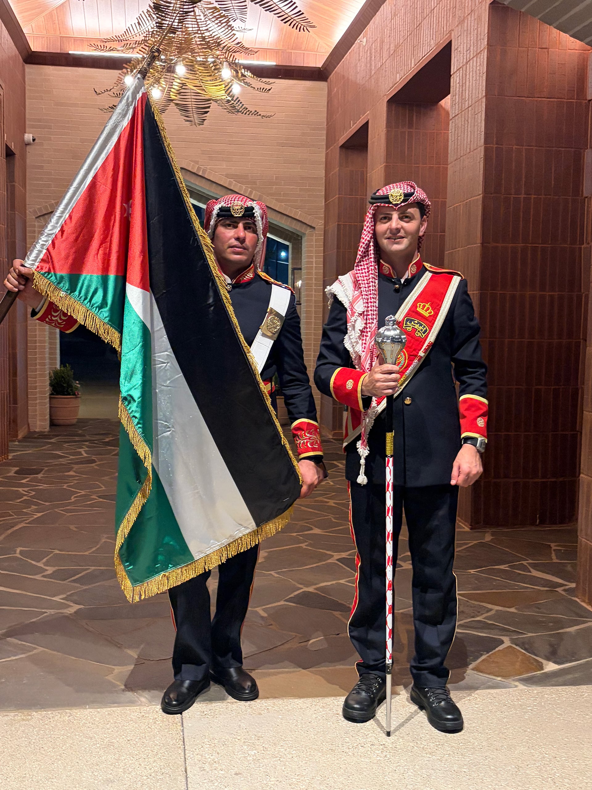 Dos integrantes de la banda militar de Jordania posan con la bandera nacional y un bastón ceremonial en un espacio interior.