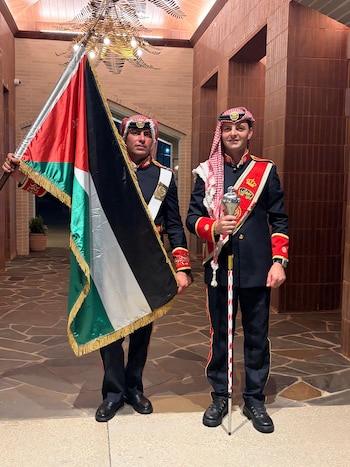 Dos hombres con uniforme militar jordano y tocados. Uno sostiene la bandera de Jordania. El otro sujeta un bastón ceremonial. Fondo de ladrillos