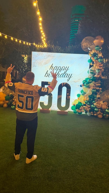 Un hombre de espaldas con una camiseta que dice Francesco 50 posa frente a un letrero de cumpleaños y un arreglo de globos