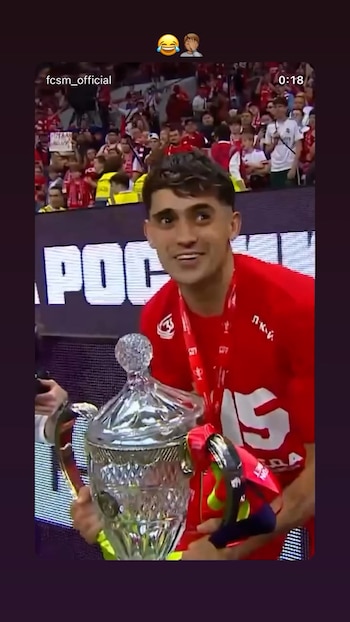 Futbolista sonriente con camiseta roja sostiene un trofeo de cristal con la tapa desprendida, entre una multitud de aficionados vitoreando
