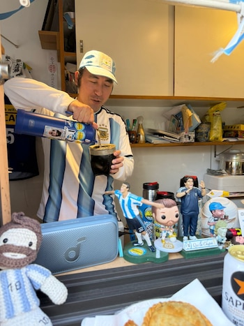 Hombre con camiseta de Argentina vierte mate en un recipiente negro. En el mostrador, figuras de fútbol, un peluche, un parlante y una empanada