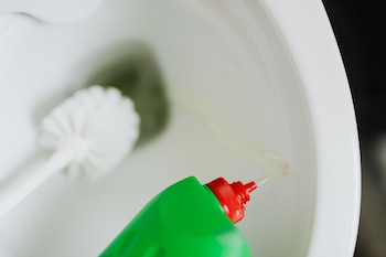 Primer plano de un inodoro blanco con un cepillo en el interior. Una mano vierte líquido limpiador de una botella verde con tapa roja