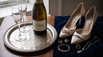 Bandeja de plata grabada, botella de champagne, dos copas, zapatos de tacón beige, anillos, pulsera, collar con brillantes sobre terciopelo azul.