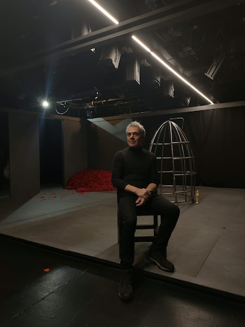 Un hombre vestido de negro está sentado en una silla en un escenario oscuro con una jaula metálica y pétalos rojos al fondo