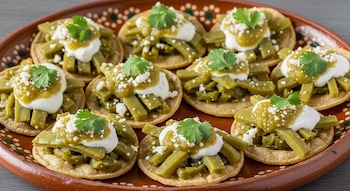Varios sopes mini en un plato de barro, cada uno cubierto con tiras de nopales cocidos, crema blanca, salsa verde, queso desmoronado y una ramita de cilantro fresco.