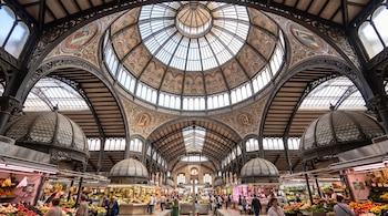 Una cúpula de cristal, hierro y cerámica sobre la nave de un mercado con puestos de frutas, verduras y personas.