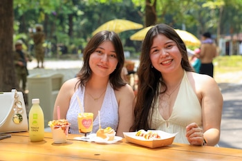 Dos mujeres jóvenes sonríen directamente a la cámara sentadas en una mesa de madera con un batido de mango, una bebida de mango limón y acompañamientos
