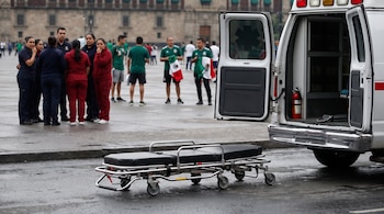 Ambulancia blanca con puertas abiertas, camilla médica en asfalto mojado. Grupo de paramédicos y aficionados de la Selección Mexicana con banderas. Edificio.