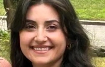 Primer plano de una mujer joven sonriendo, con cabello oscuro y piel clara. Se ve parte de vegetación verde borrosa de fondo