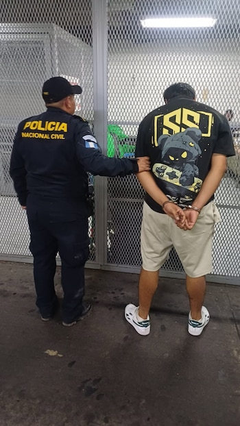 Hombre con uniforme de Policía Nacional Civil de Guatemala y gorra negra sujeta a un hombre de espaldas con camiseta negra, shorts beige y esposas