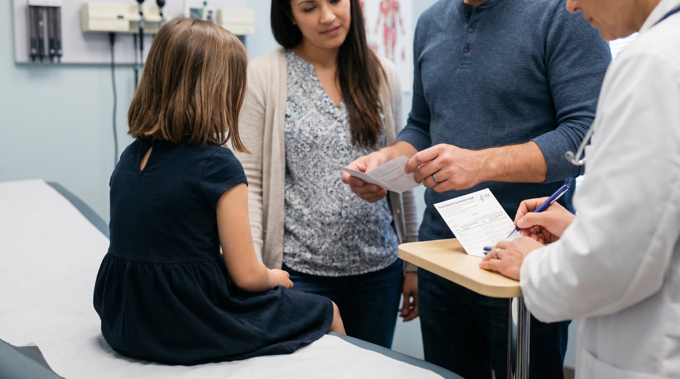 Un médico firma el carnet de vacunación de una niña, quien está sentada en la camilla acompañada por sus padres durante una consulta de rutina. (Imagen Ilustrativa Infobae)