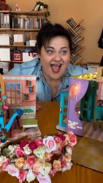 Mirta Wons sonríe detrás de una mesa con maquetas de habitaciones en miniatura y una guirnalda de flores de colores