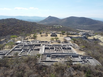 Vista aérea de las ruinas de Xochicalco, con estructuras de piedra y terrazas escalonadas, rodeadas de vegetación seca y montañas en el horizonte bajo un cielo azul