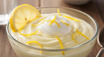 Un cuenco de vidrio con postre cremoso de color amarillo claro, una rodaja de limón, un montículo de crema blanca y tiras de ralladura amarilla.
