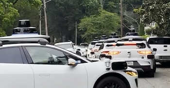 Una fila de coches blancos autónomos con equipos de sensores negros y azules en sus techos, estacionados o en movimiento en una calle arbolada