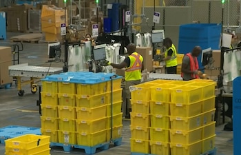 Empleados en un almacén de Amazon procesan paquetes y cajas en estaciones de trabajo, con grandes pilas de contenedores amarillos y azules visibles en el suelo