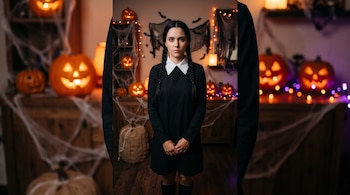Mujer con vestido negro, cuello blanco y dos trenzas de pie frente a calabazas decoradas con caras, telarañas artificiales y luces.