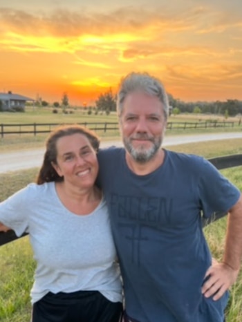 Mujer y hombre sonrientes abrazados. Detrás, un atardecer naranja con nubes, un campo verde, una valla de madera y una casa en la distancia