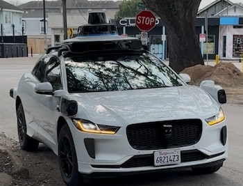 Un coche autónomo Waymo blanco, con múltiples sensores en el techo y los laterales, está estacionado junto a una acera en una calle urbana