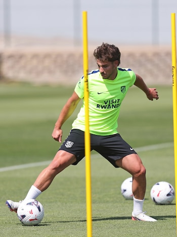 Jugador de fútbol masculino con camiseta verde y pantalón negro con logo del Atlético de Madrid, entrenando en un campo de césped con balones y postes amarillos