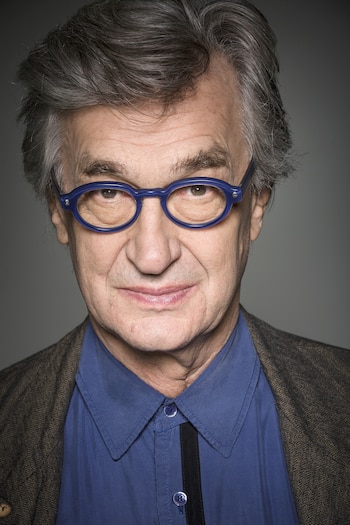 Retrato de primer plano de un hombre con anteojos de marco azul, camisa azul y saco sobre un fondo de tono gris