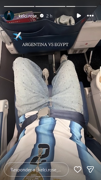 Persona sentada en un avión con jeans y camiseta de la selección argentina. Tiene el cinturón de seguridad abrochado. La pantalla frontal indica 'ARGENTINA VS EGYPT'