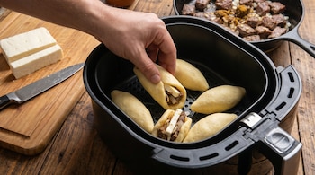 Una mano introduce carimañolas crudas, rellenas de carne y queso, en la cesta de una freidora de aire. En la mesa hay queso y una sartén con carne.