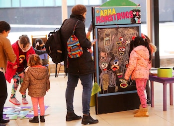 Varias personas, incluidos niños y adultos, interactúan con una pizarra que exhibe dibujos de monstruos y un letrero que dice 'ARMA MONSTRUOS'