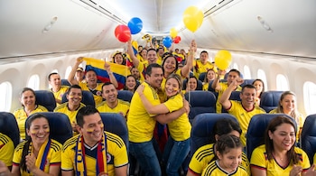 Interior de un avión con decenas de hinchas colombianos sonriendo y celebrando, vistiendo camisetas amarillas y con banderas y globos.