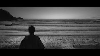 Figura de espaldas con moño, de pie en playa de arena oscura con rocas y olas. Colinas en el horizonte. Imagen monocromática