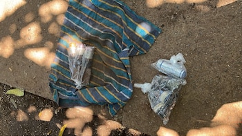 Objetos envueltos en plástico transparente, incluyendo un cilindro blanco con 'GL-202', y un paño a rayas azul y amarillo, sobre suelo terroso con luz solar