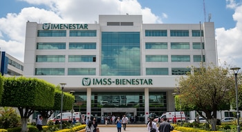Fachada principal del edificio del Hospital IMSS-BIENESTAR, de color blanco con grandes ventanales de cristal, un área ajardinada frontal y personas en la entrada.