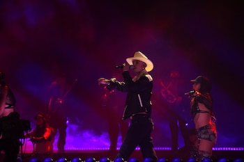 Un cantante con sombrero blanco y micrófono actúa en un escenario iluminado con luces moradas junto a bailarines y músicos