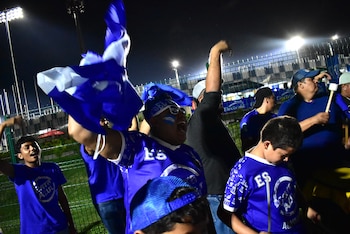 Personas con camisetas azules con la sigla ES ondean una bandera blanca y azul de noche junto a una cerca de alambre