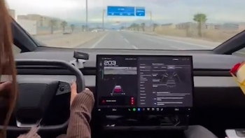 Interior de un automóvil con una persona al volante. Pantalla central muestra 203 km/h. Se observa una carretera y un cartel vial azul