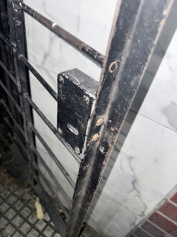 Primer plano de una reja de seguridad de metal negro con un mecanismo de cerradura. Se aprecian detalles de corrosión y el marco de metal, con una pared clara detrás