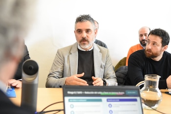Un hombre con barba y chaqueta gris habla en una mesa de reuniones junto a un ordenador portátil y una jarra de agua