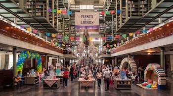 Interior de una biblioteca con estanterías elevadas, un esqueleto suspendido, adornos de colores, visitantes y un cartel de Fiesta de los Libros.