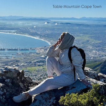 Mujer sentada en una roca en la cima de una montaña, con un paisaje de ciudad y mar de fondo, bajo un cielo azul