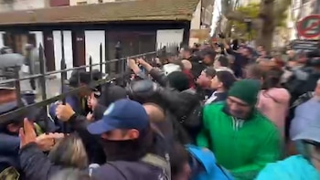 Multitud de personas se congregan en una calle, algunas empujan una valla metálica negra. Se ven edificios al fondo y árboles