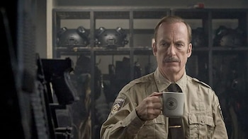 Un hombre con bigote, de uniforme de sheriff beige, sostiene una taza gris con un emblema, mirando fijamente. Estantes con equipo visible detrás