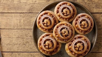 Vista cenital de seis roles de manzana y canela con azúcar glas en una bandeja metálica oscura. El relleno visible contiene trozos de manzana y especias