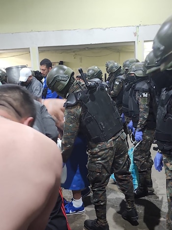 Un grupo de infantes con uniformes de camuflaje, cascos y chalecos antibalas dentro de un edificio. Varios civiles son visibles, uno de ellos con la espalda desnuda