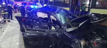Coche negro con daños severos, parabrisas roto, airbags desplegados. Bomberos y policías en la escena, cerca de un camión de bomberos